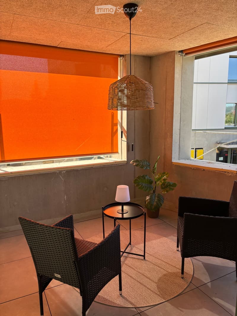 Balcony with two wicker chairs, a small round table with a lamp, and a potted plant. Features large windows and modern decor.