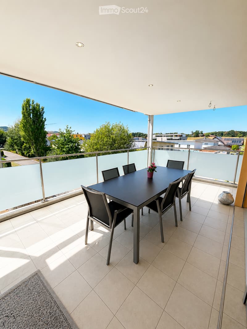 Balcony with a dining table, six chairs, and a flower pot, overlooking a scenic view of trees and buildings.