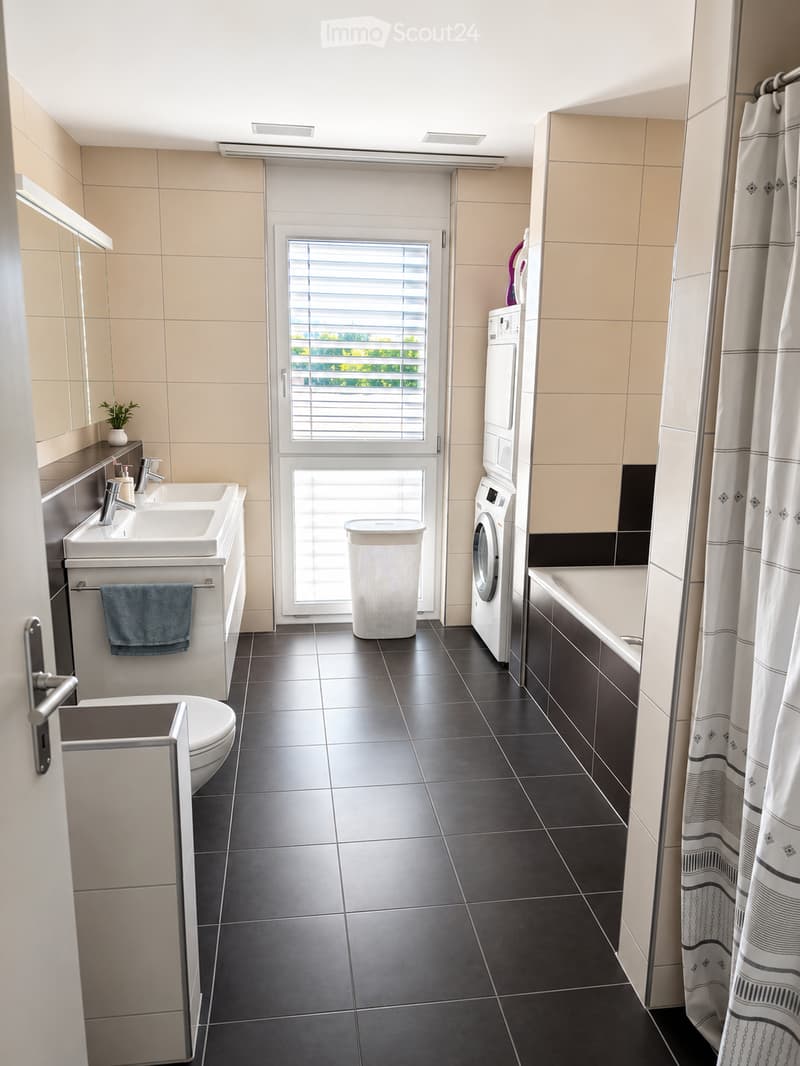Modern bathroom with double sink, toilet, bathtub, washing machine, and a window with blinds. Features tiled walls and floor, a shower curtain, and a small plant on the sink counter.