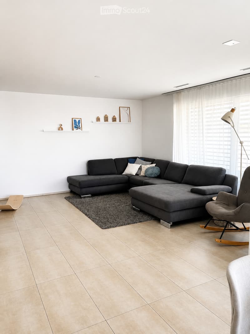 Modern living room with a grey sectional sofa, ottoman, armchair, floor lamp, and decorative shelf with picture frames and small house models.