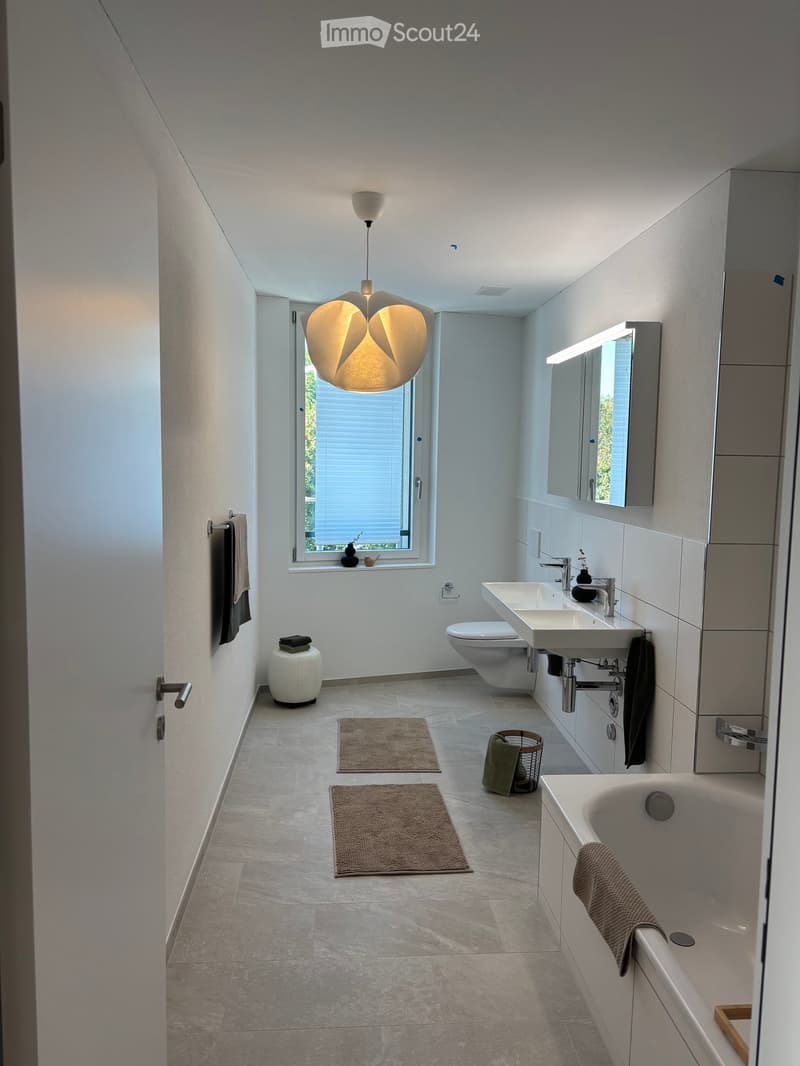Modern bathroom with a bathtub, sink, toilet, large mirror, and a hanging light fixture. Includes towels, a small stool, and rugs on the floor.