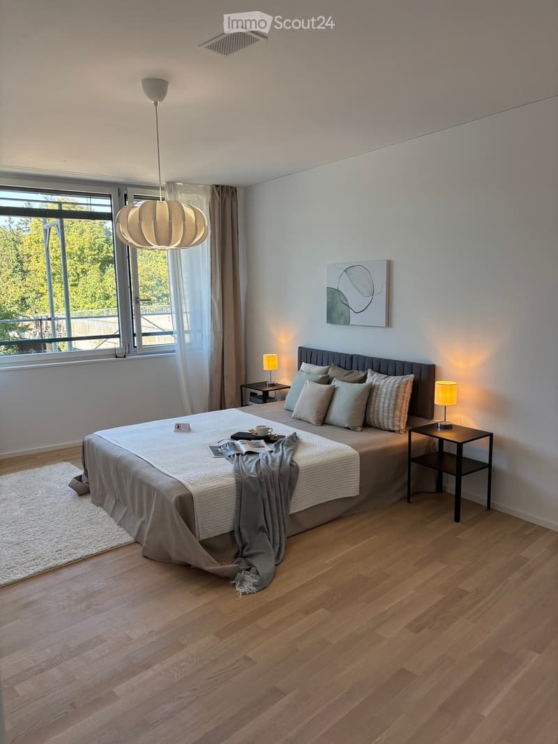 Modern bedroom with wooden flooring, large bed with pillows and blankets, nightstands with table lamps, pendant light, artwork on wall, and large window with view of greenery.