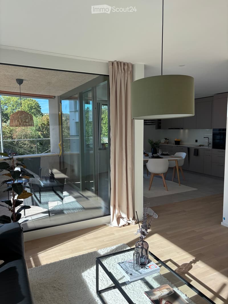 Modern living room with a black leather sofa, glass coffee table, and a dining area with white chairs and a wooden table. The space features large windows with a view, a balcony, and a kitchen area with cabinets and a sink.