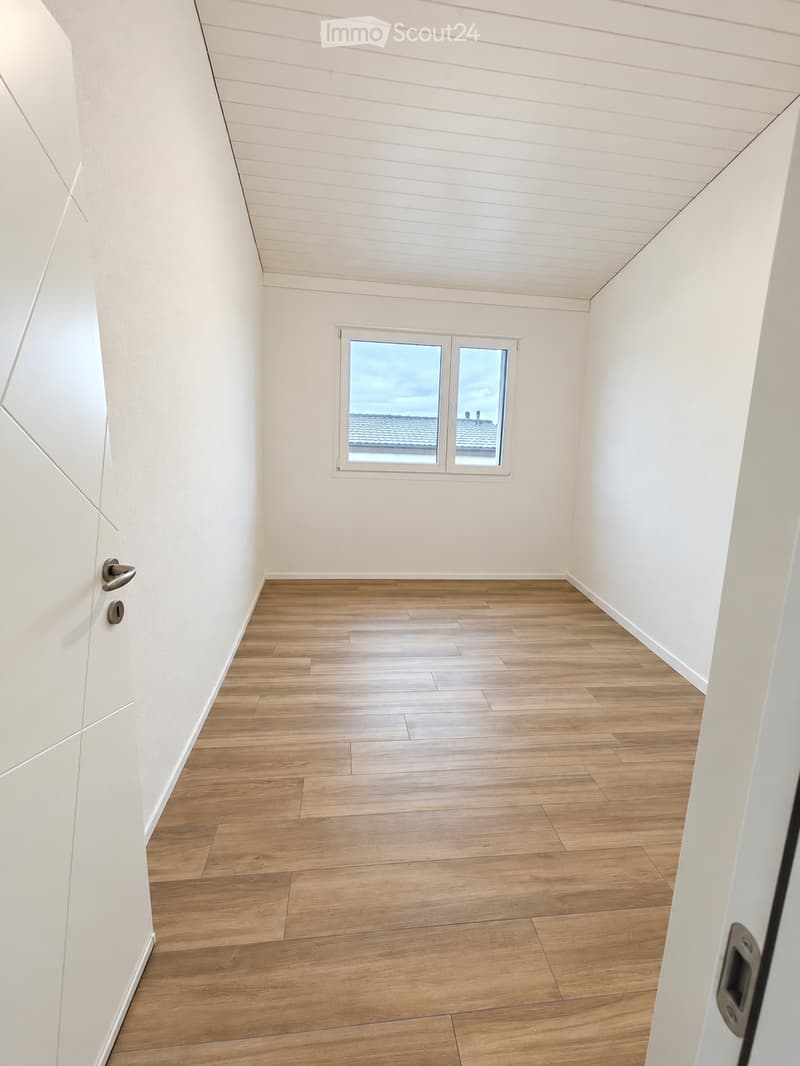 Empty room with wooden flooring, white walls, and a window offering a view of a roof and chimney.