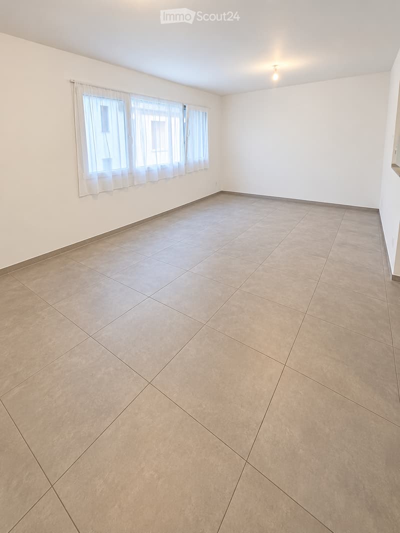 Empty room with tiled floor and white walls, featuring large windows with sheer curtains and a single ceiling light fixture.