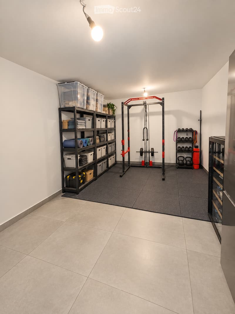 Well-lit home gym with a multi-tiered black shelving unit holding storage bins, towels, and exercise mats. A red and black multi-functional exercise machine is positioned centrally, accompanied by a rack of dumbbells, kettlebells, and a water bottle. A sm