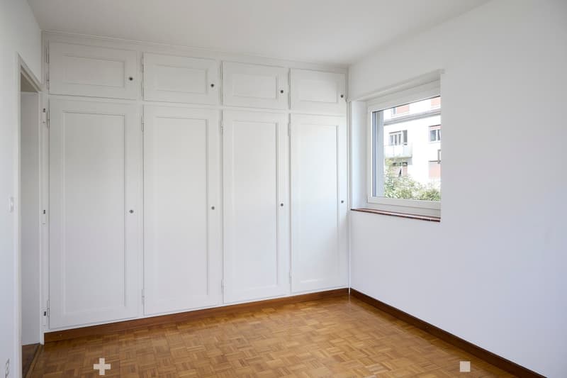 Empty room with white walls, wooden floor, built-in white cabinets, and a window with a view of a building and greenery outside.