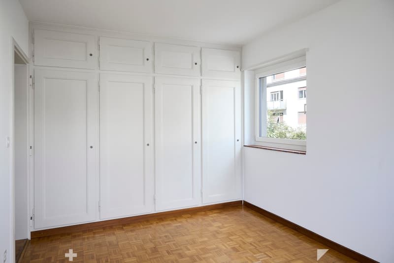 Empty room with white walls, wooden floor, built-in white cabinets, and a window with a view of a building and greenery outside.