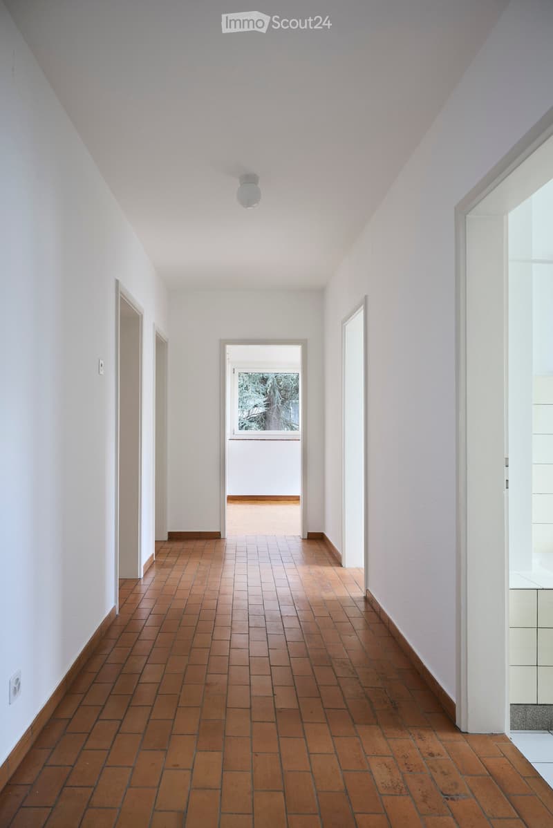 A long, narrow hallway with white walls, wooden tiled flooring, and several doors leading to other rooms. The ceiling is white with a single light fixture.