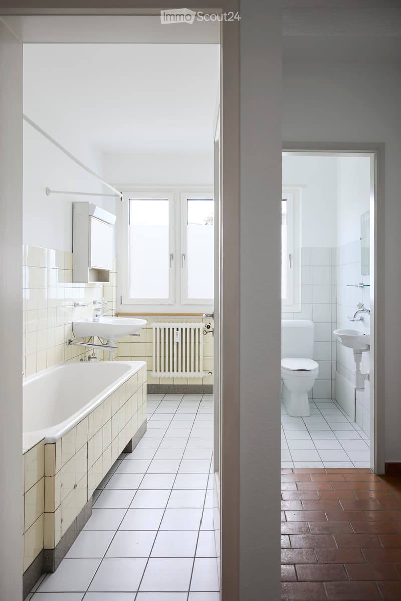 Two adjacent bathrooms with white tiles, a bathtub, sink, toilet, and radiator. One bathroom has a window, while the other has a mirror and cabinet.