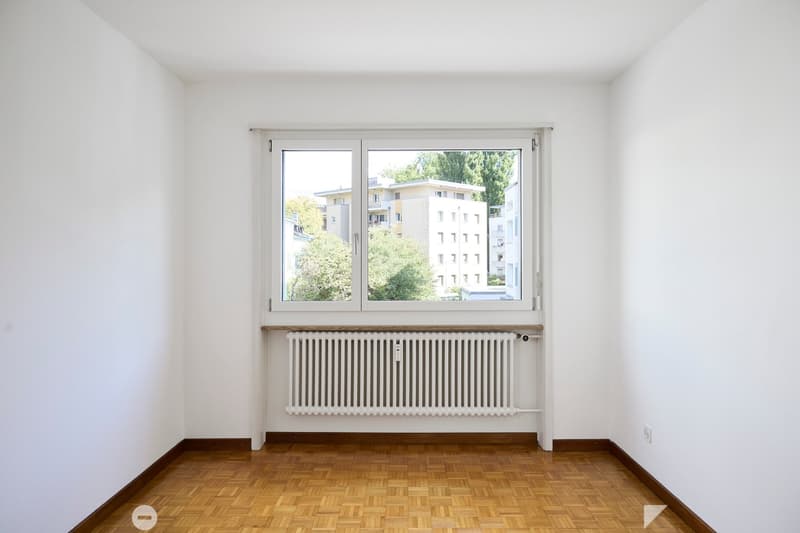 Empty room with white walls, wooden floor, large window with radiator below, and visible electrical outlet on the wall.