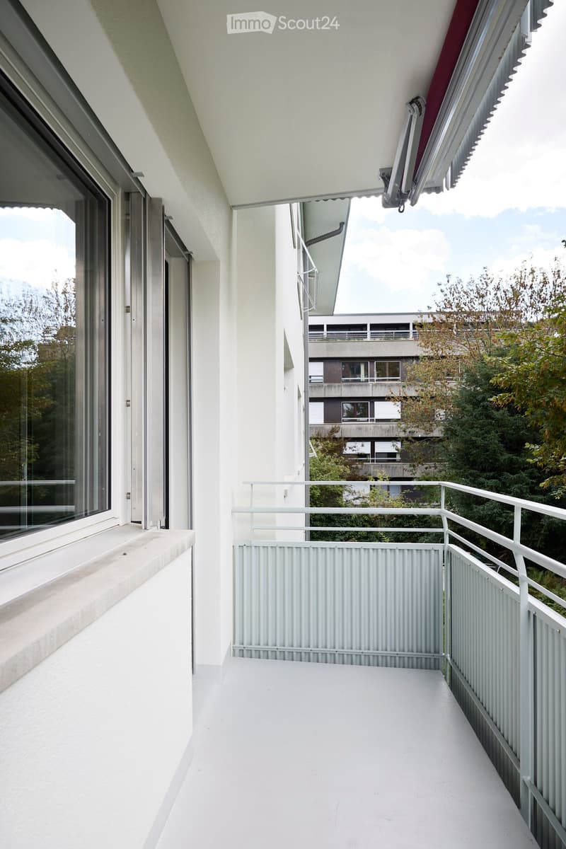 Modern balcony with white flooring, metal railing, and large windows offering a view of the surrounding greenery and buildings.