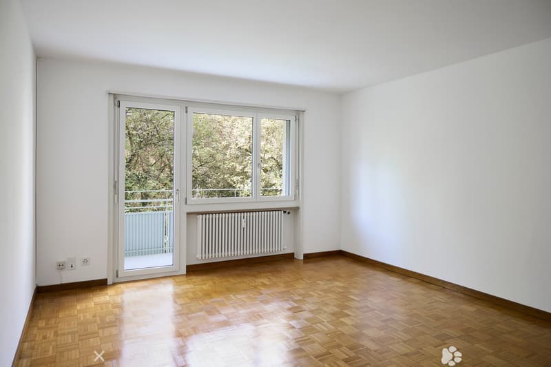 Empty room with white walls, wooden floor, large window with view of trees, radiator, and sliding glass door leading to a balcony.