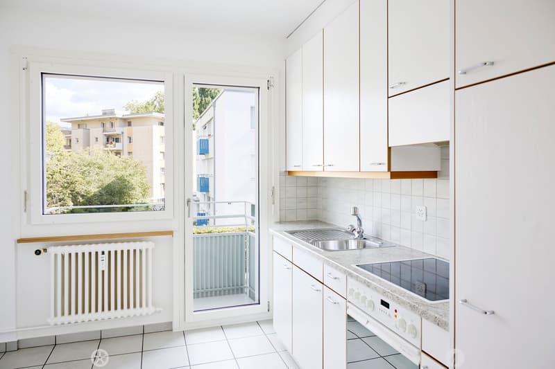 Modern kitchen with white cabinets, stainless steel sink, oven, and a window overlooking a residential area.
