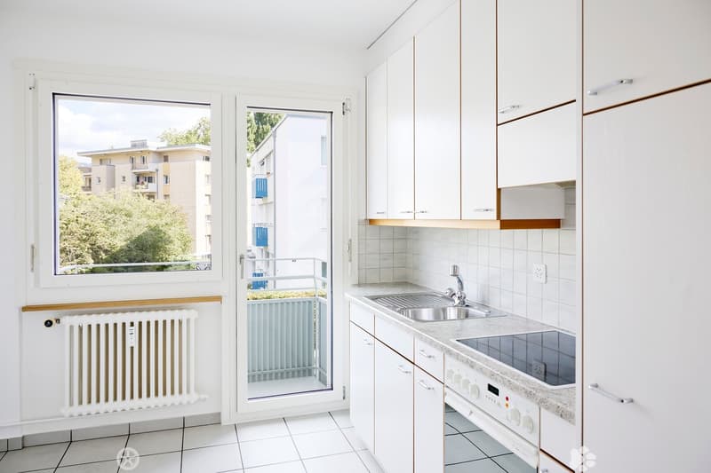 Modern kitchen with white cabinets, stainless steel sink, oven, and a window overlooking a residential area.
