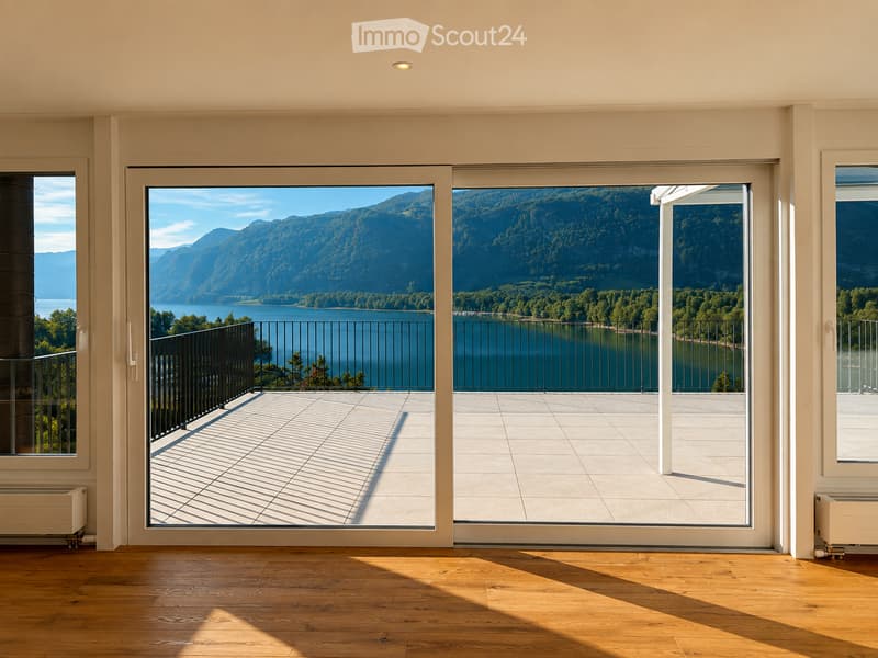 Bright, unfurnished room with large sliding glass doors opening to a balcony overlooking a scenic lake and mountains. Wooden flooring and white walls.