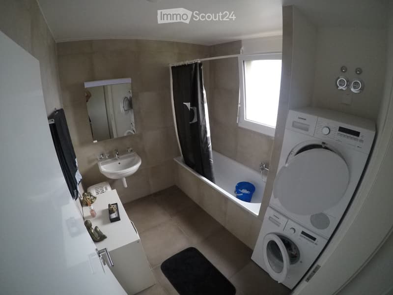 Modern bathroom with a sink, mirror, bathtub, washing machine, and shower curtain. The space is well-lit with a window and includes a small stool and a light switch.