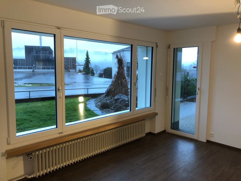 Empty room with wooden flooring, large windows, radiator, and a sliding glass door leading to a small outdoor area.