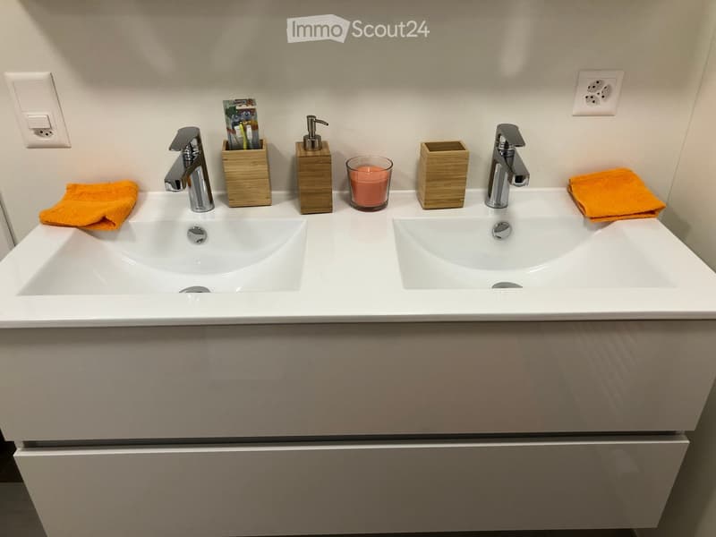 Modern bathroom with a double sink vanity, two faucets, two towels, a soap dispenser, a toothbrush holder, and a candle on the counter.