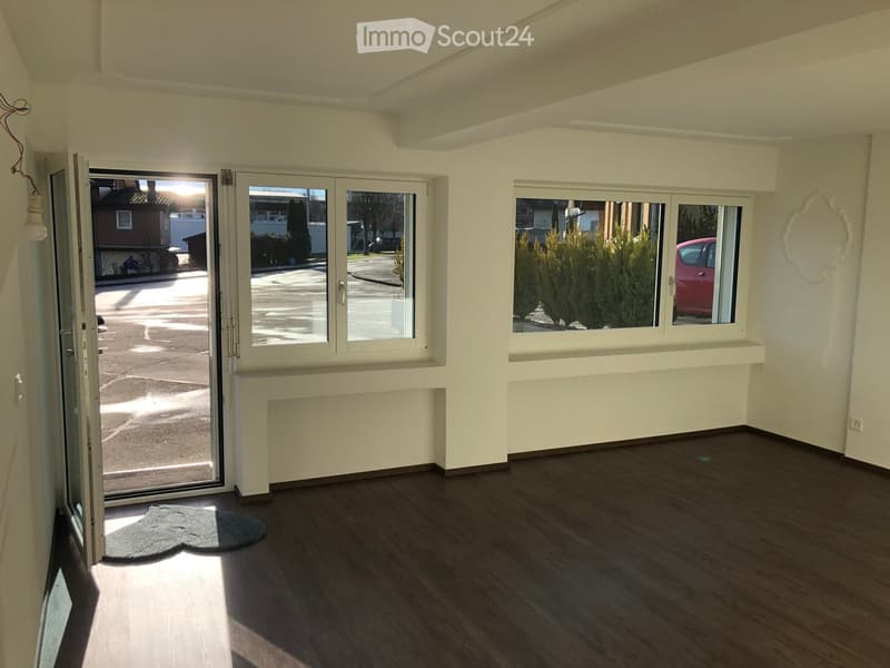 Empty room with wooden flooring, large windows, and a sliding glass door leading outside. The room has white walls and a small rug near the entrance.
