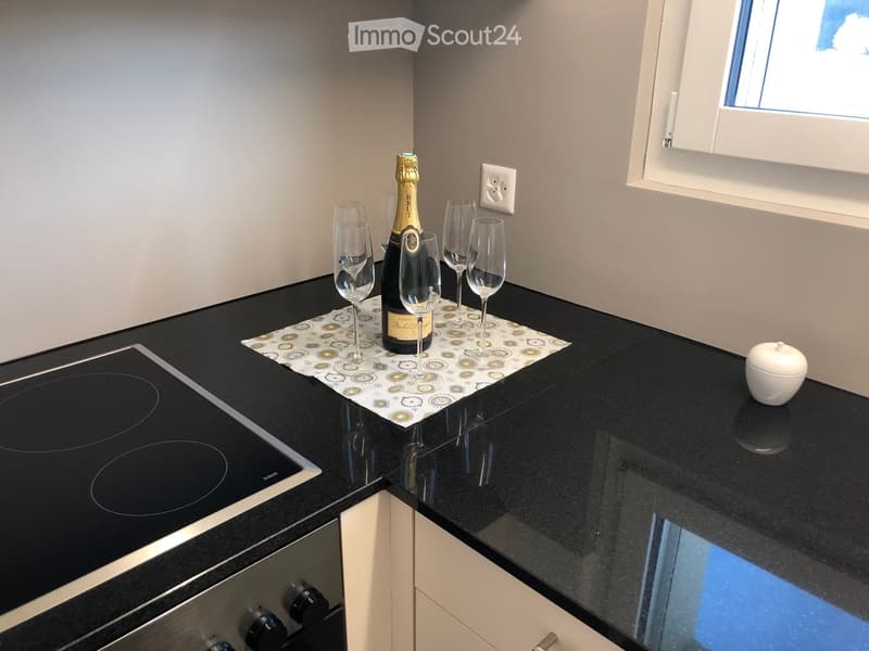 Modern kitchen with black granite countertops, electric stove, and wine glasses and bottle on a mat.