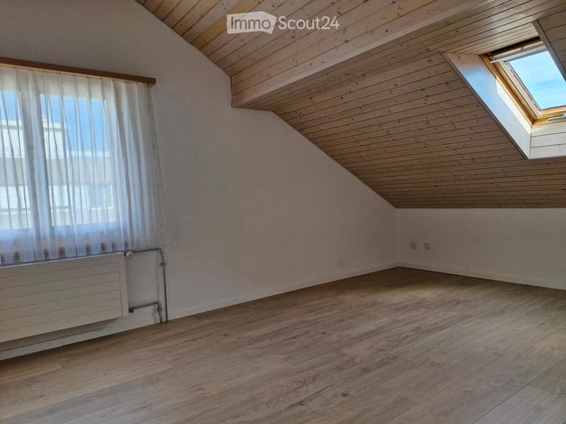 Empty attic room with wooden floors, wooden slanted ceiling, white walls, and a skylight window. There is a radiator on one wall and a curtain covering a window.
