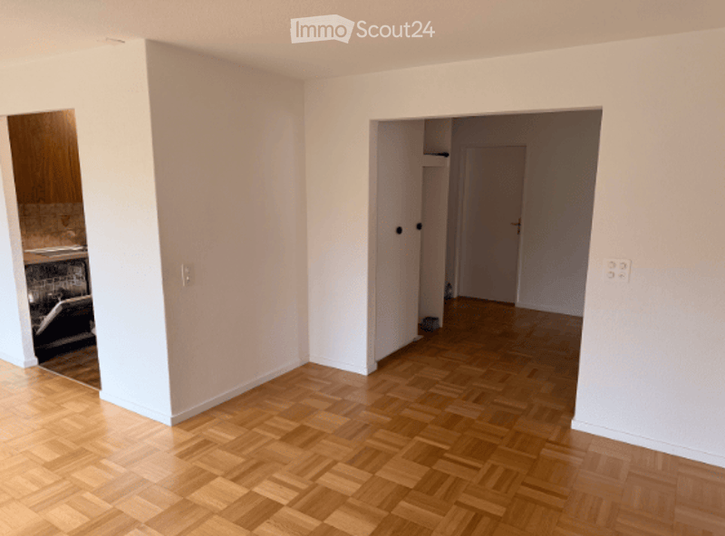 Empty room with light-colored walls and a wooden parquet floor. There is a doorway leading to another room and a visible kitchen area in the background.