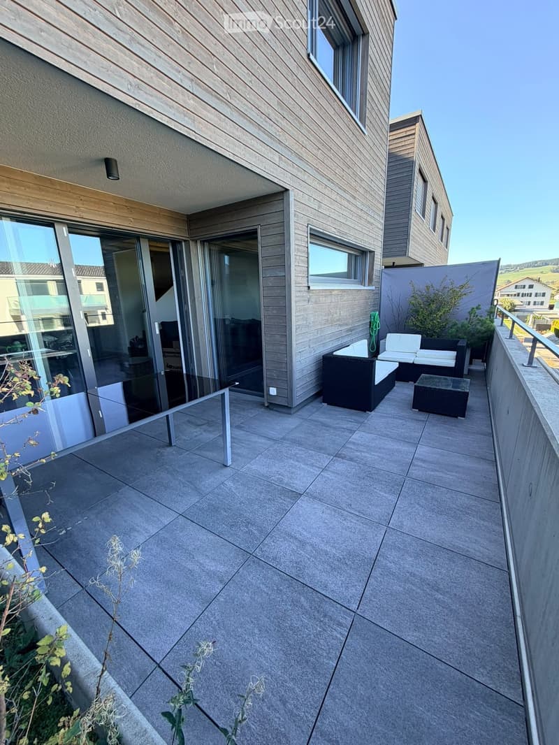 Modern balcony with grey tiled floor, black wicker furniture including a sofa, table, and chairs, glass sliding doors leading to the interior, and a metal railing.