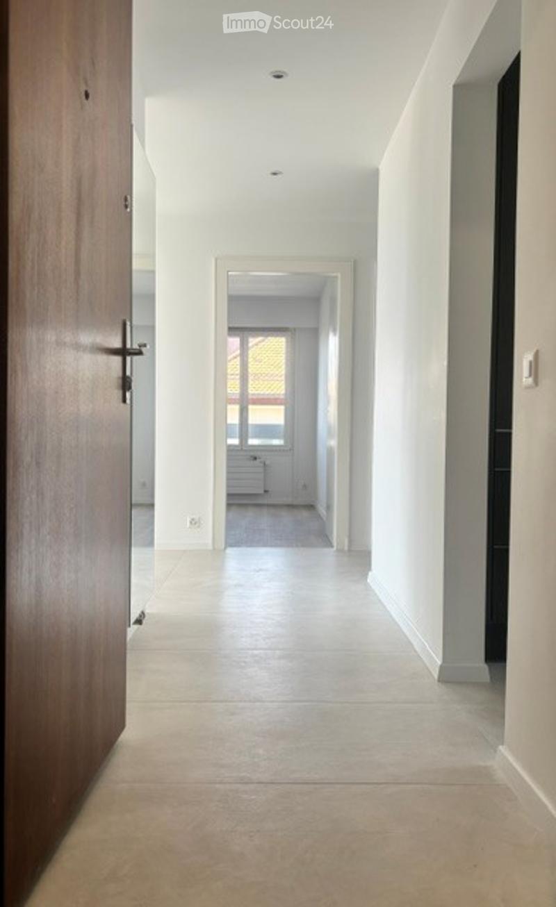 Empty hallway with white walls, tiled floor, and a wooden door on the left. A distant room with a window and radiator is visible at the end.