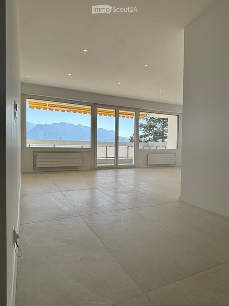 Bright, empty room with large sliding glass doors leading to a balcony, offering a scenic mountain view. The room features a tiled floor and white walls.