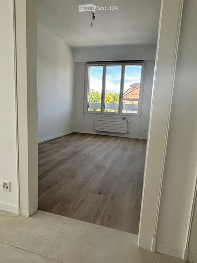 Empty room with wooden flooring, white walls, and a large window with a view of greenery and a neighboring building.