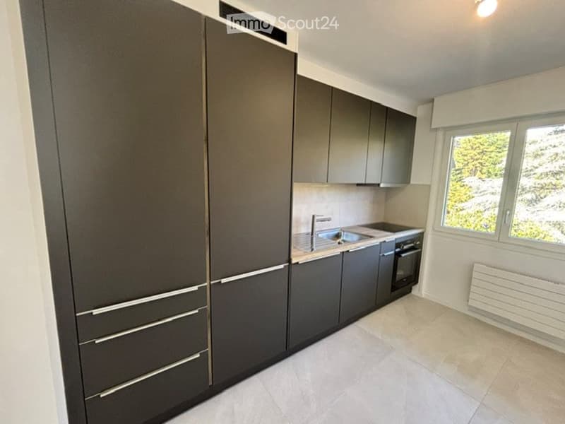 Modern kitchen with dark grey cabinetry, stainless steel sink, and oven. Large window offers a view of greenery outside.