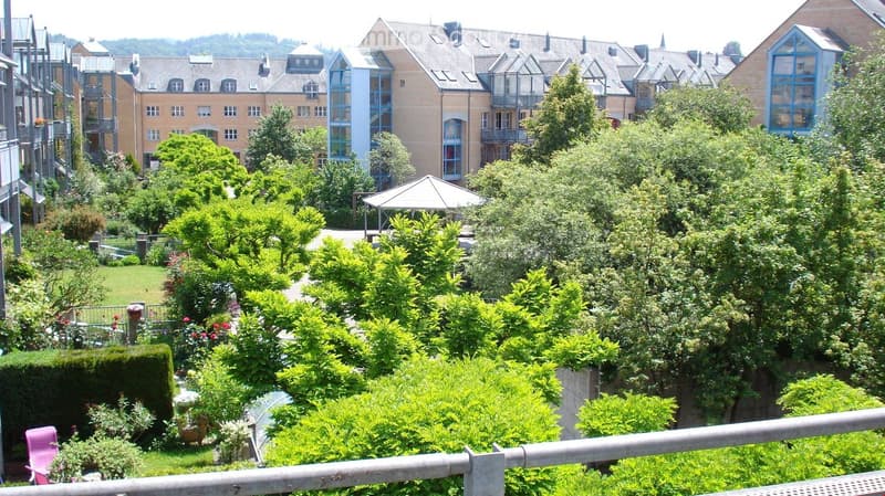 View from a balcony or terrace showing a landscaped garden area with trees, shrubs, and a small gazebo, with residential buildings in the background.