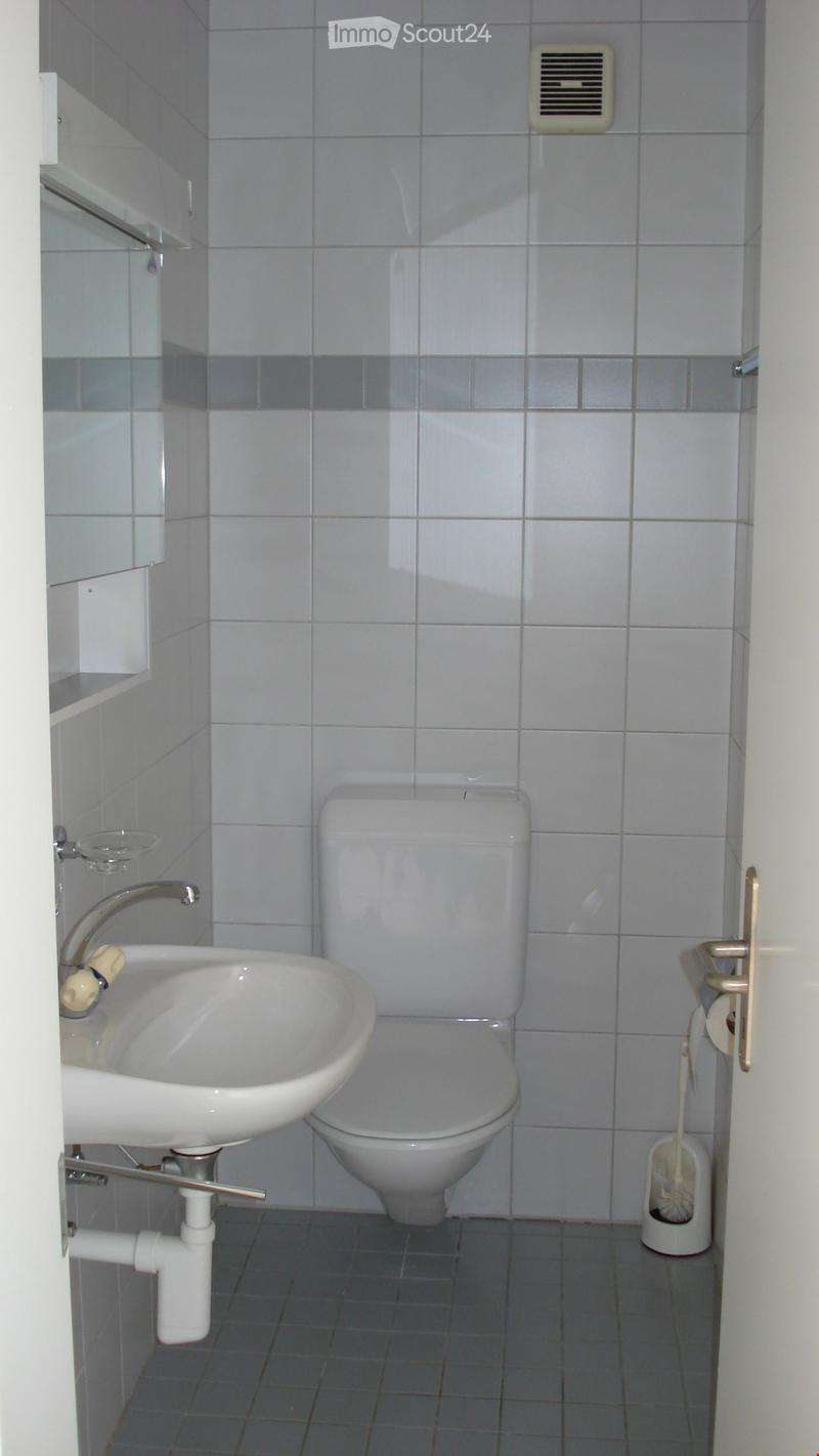 A small bathroom with white tiled walls and floor, featuring a toilet, a sink with a faucet, a toilet paper holder, and a toilet brush holder.