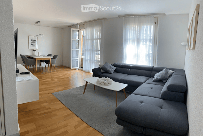 Modern living and dining area with a grey sectional sofa, white coffee table, dining table with four chairs, and a TV on a white stand. The space features wooden flooring, large windows with sheer curtains, and a grey rug.