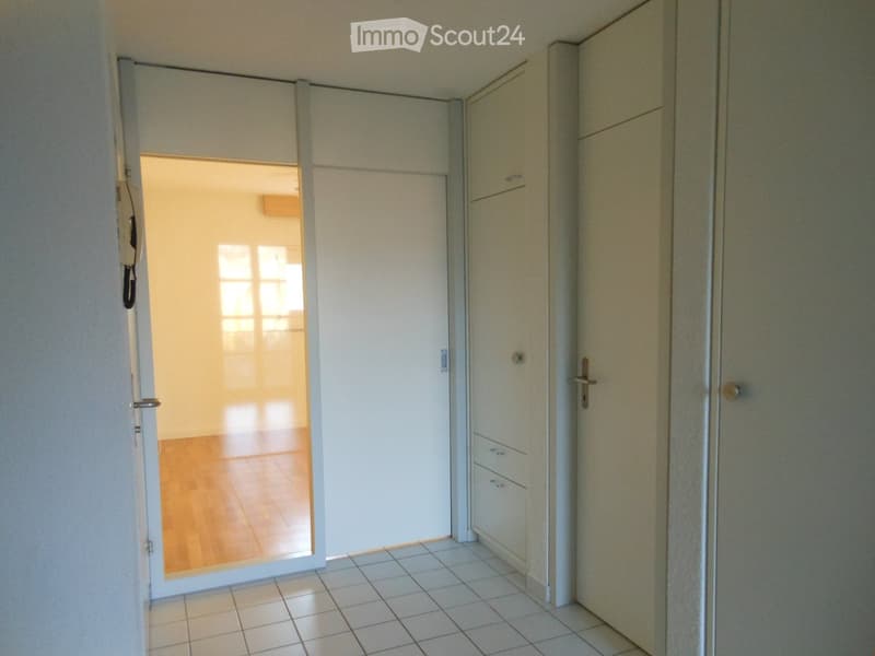 A narrow hallway with tiled flooring, white walls, and a built-in storage unit with drawers and cabinets. An open door leads to a room with wooden flooring and large windows.