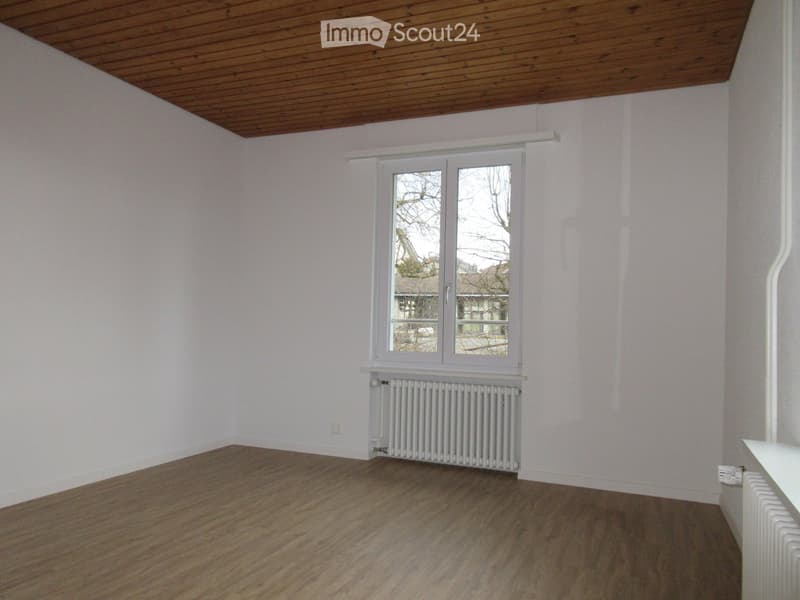 Empty room with wooden flooring, white walls, and a large window with a radiator below it.