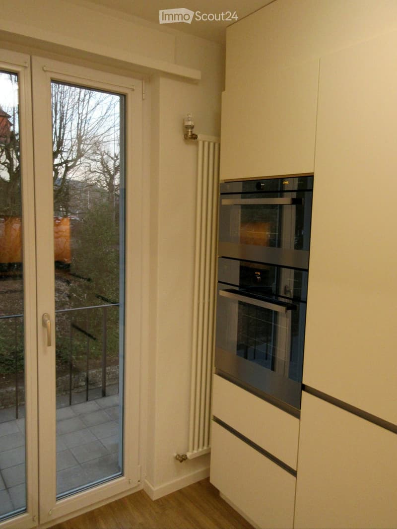 Modern kitchen with light-colored cabinets, built-in oven, and a large window leading to a tiled balcony area.