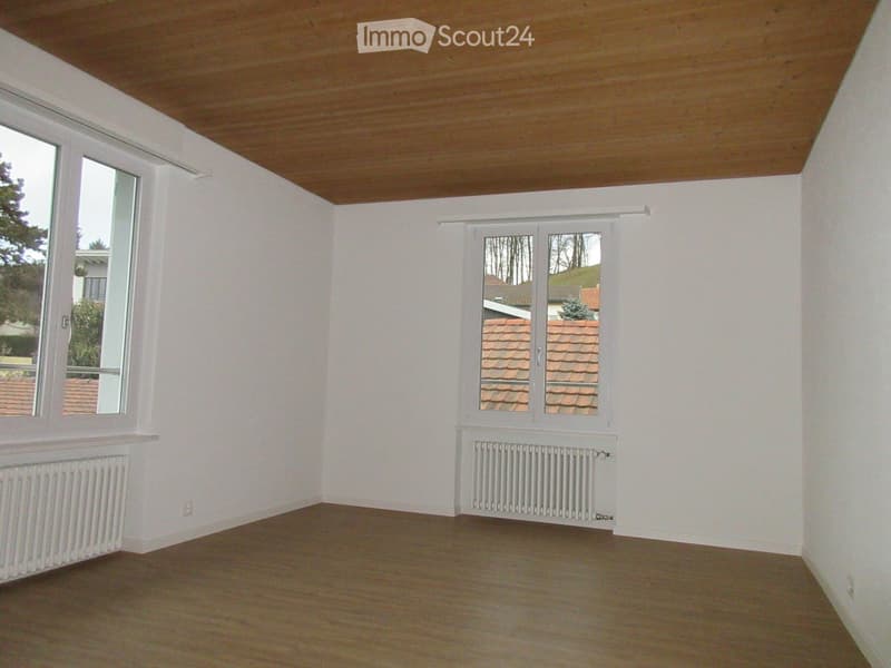 Empty room with white walls, wooden floor, and large windows showing a view of a roof and trees outside.