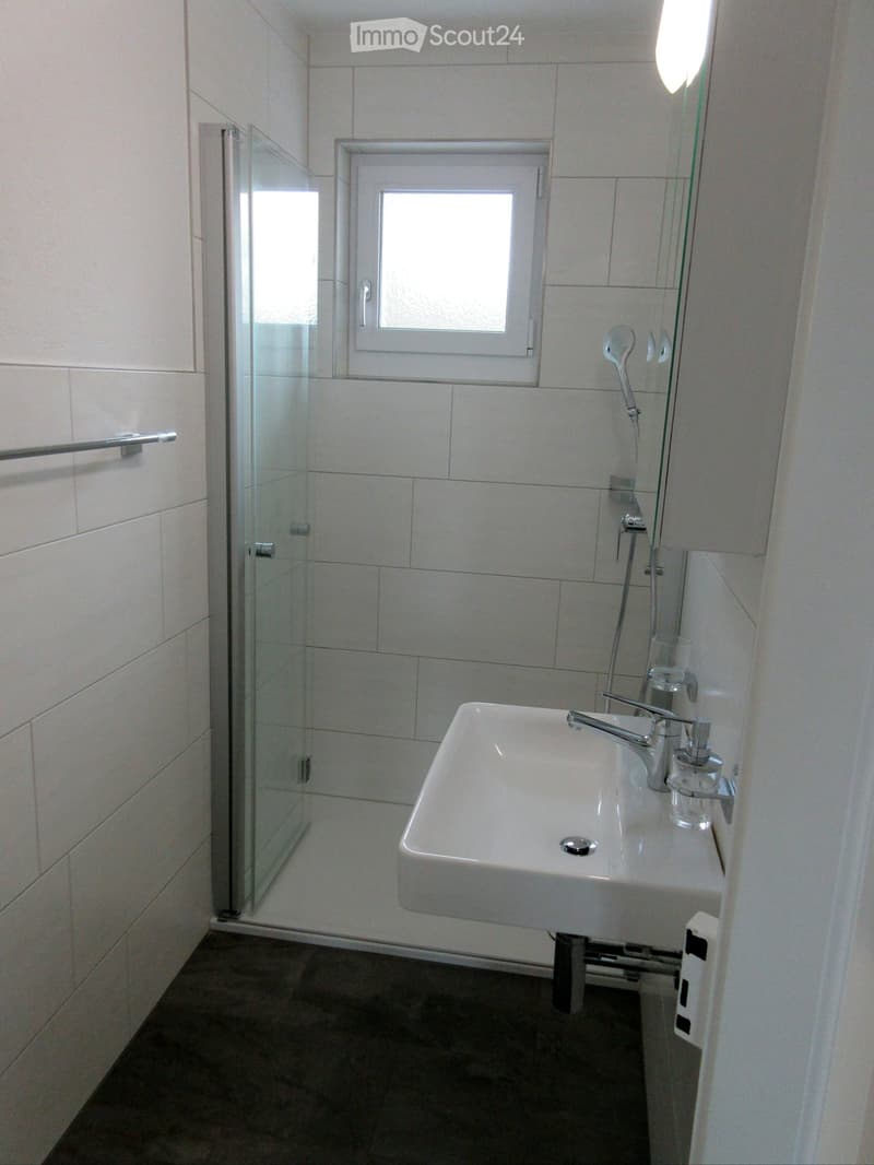 Modern bathroom with white tiled walls, a white sink with a chrome faucet, a glass-enclosed shower, and a window above the sink.