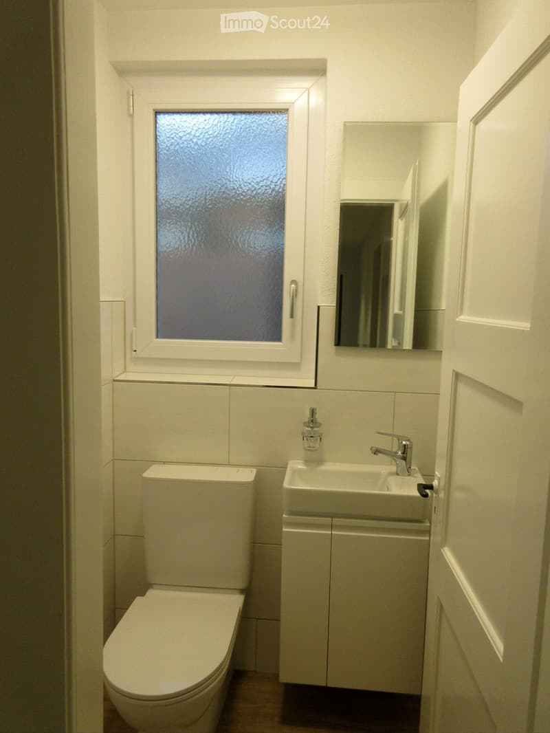 Modern bathroom with white tiles, a toilet, a sink with a modern faucet, a mirror, and a frosted window above the sink.
