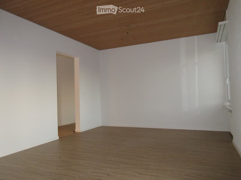 Empty room with white walls, wooden floor, and a wooden ceiling. There is a doorway leading to another room.