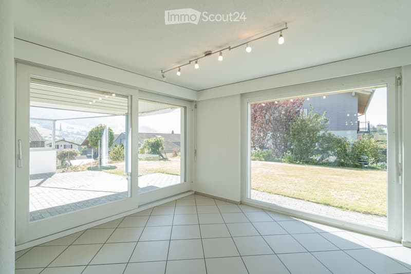 Bright, empty room with large glass sliding doors leading to a garden, white tiled floor, and recessed lighting track on the ceiling.