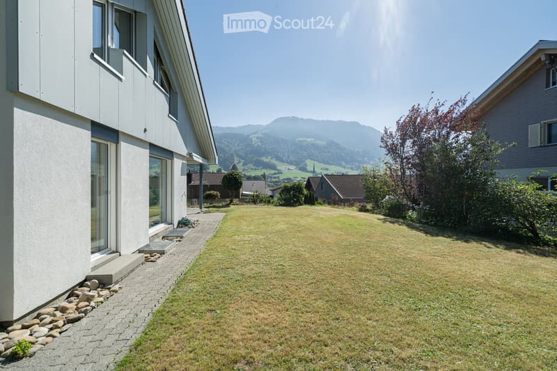 Modern two-story house with white exterior, large windows, and a well-maintained lawn in front. The house features a raised ground floor and a scenic mountain view in the background.