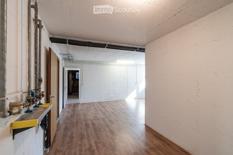 Empty hallway with wooden flooring, white walls, and exposed pipes on one side. Natural light streams in from the far end, revealing an open space beyond.