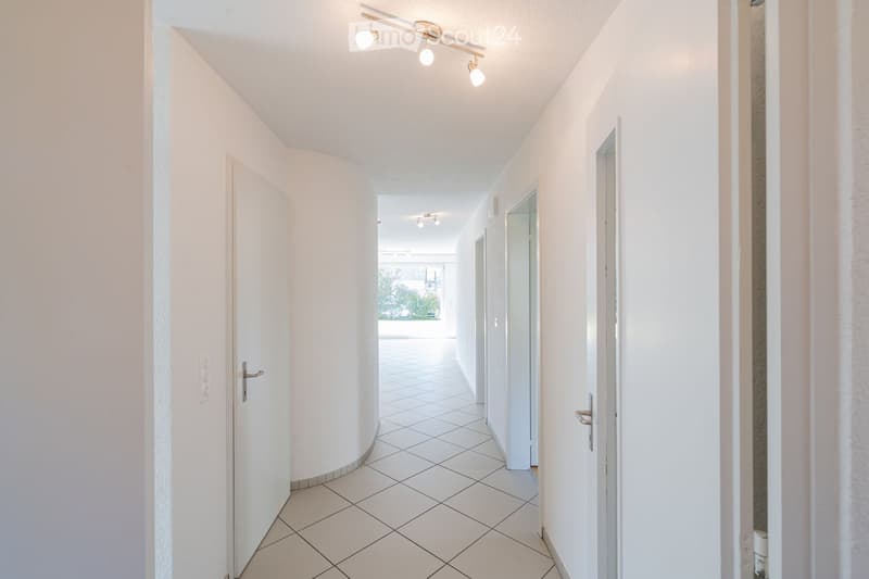 A clean, modern hallway with white tiled flooring, white walls, and recessed lighting. Doors are visible on both sides, leading to other rooms.
