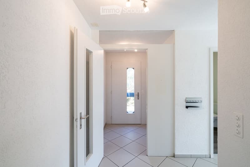 A clean, modern hallway with white walls, tiled flooring, and recessed lighting. Features include a white door with a small window, a mail slot, and a light switch on the wall.