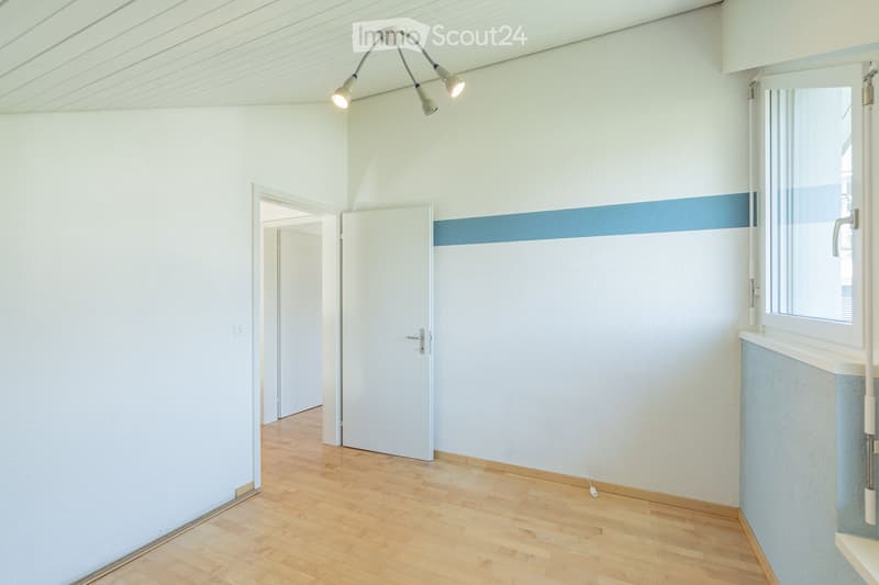 Empty room with light-colored walls, wooden flooring, and a ceiling light fixture. There is a window with a blue accent strip and a door leading to another room.