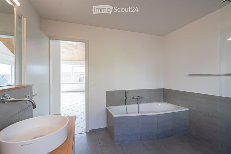 Modern bathroom with a white sink, bathtub, and a large mirror. The room features grey tiles and a glass shower enclosure.