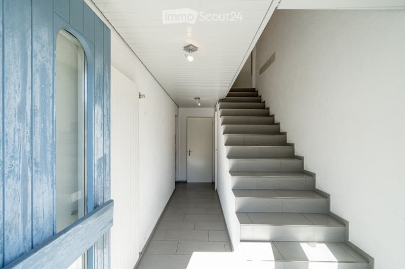 Modern hallway with white walls, tiled floor, and a staircase leading upwards. Features recessed lighting and a glass-paneled door on the left.